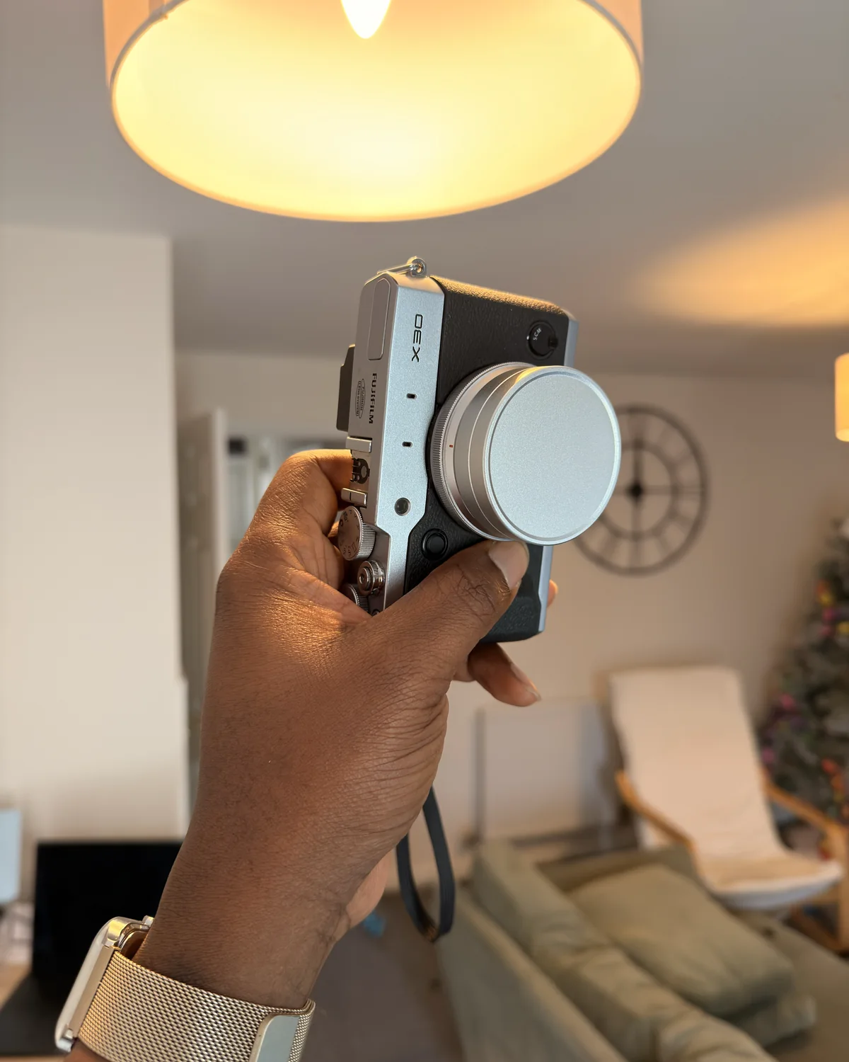 A hand holding up a silver compact camera in a living room under a warm lamp, with a wall clock and a Christmas tree behind